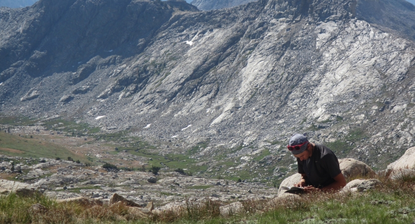 A person sits on a rock and journals. There is a mountain in the background.